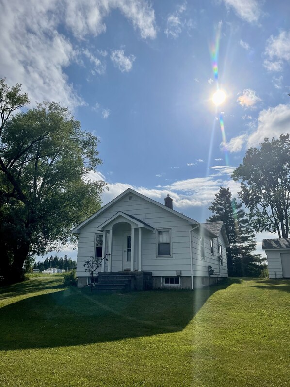 A white single-story house with a front porch, surrounded by a well-maintained lawn and trees. - Quaint Home in a Great Central Location (Rapid River)