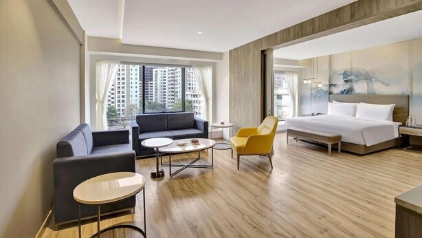 A modern hotel room with a large bed, a sofa, a yellow armchair, a small round table, and a view of high-rise buildings through the window. - Park Inn by Radisson Surat (Surat)
