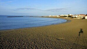 Une plage de sable avec des empreintes de pas qui mènent à l’eau, un ciel dégagé et des bâtiments en arrière-plan.