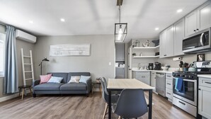A modern kitchen with white cabinets, a microwave, and an oven. There is a dining table with chairs and a gray sofa with pillows.