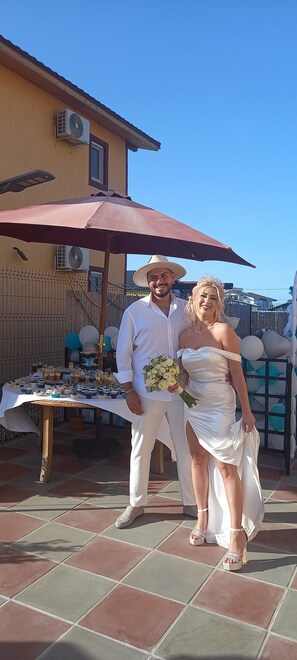A couple standing under a red umbrella in an outdoor setting with a table displaying various items in the background.