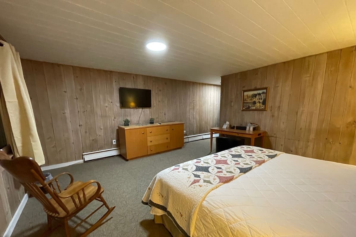 A bedroom with a wooden headboard bed, a wooden dresser, a desk with a chair, a rocking chair, a flat-screen TV mounted on the wall, and a painting on the wall.
