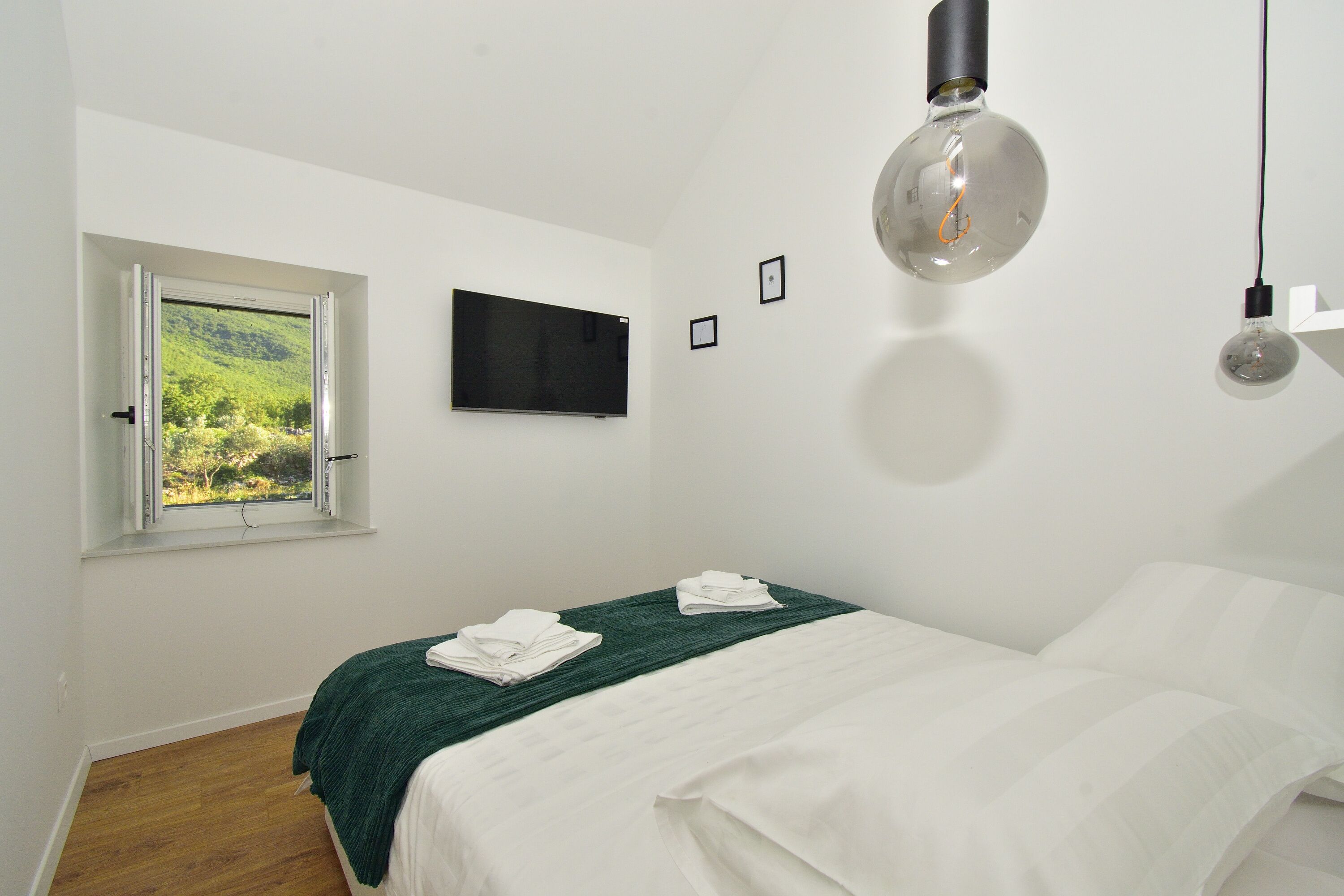 Ein Schlafzimmer mit Bett, einem Fenster mit Blick auf Grünflächen, einem Flachbildfernseher und zwei Wandleuchten.