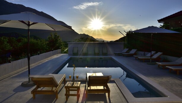 A swimming pool with lounge chairs and umbrellas, surrounded by mountains and a clear sky.