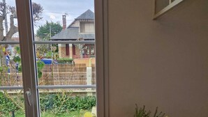 Una ventana con vistas a una zona residencial, una mesa con frutas y frutos secos, y un florero con flores.