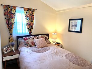 Une chambre à coucher avec un lit, une table de chevet avec une lampe, un tableau encadré au mur et une fenêtre avec des rideaux à motifs floraux.