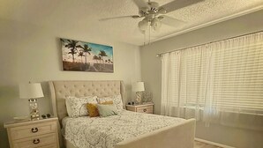 Un dormitorio con cama, mesitas de noche, ventilador de techo, un cuadro y una ventana con persianas.