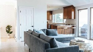 Una sala moderna con un sofá seccional gris, una mesa de centro redonda y vista a una cocina con armarios de madera y un ventanal grande.