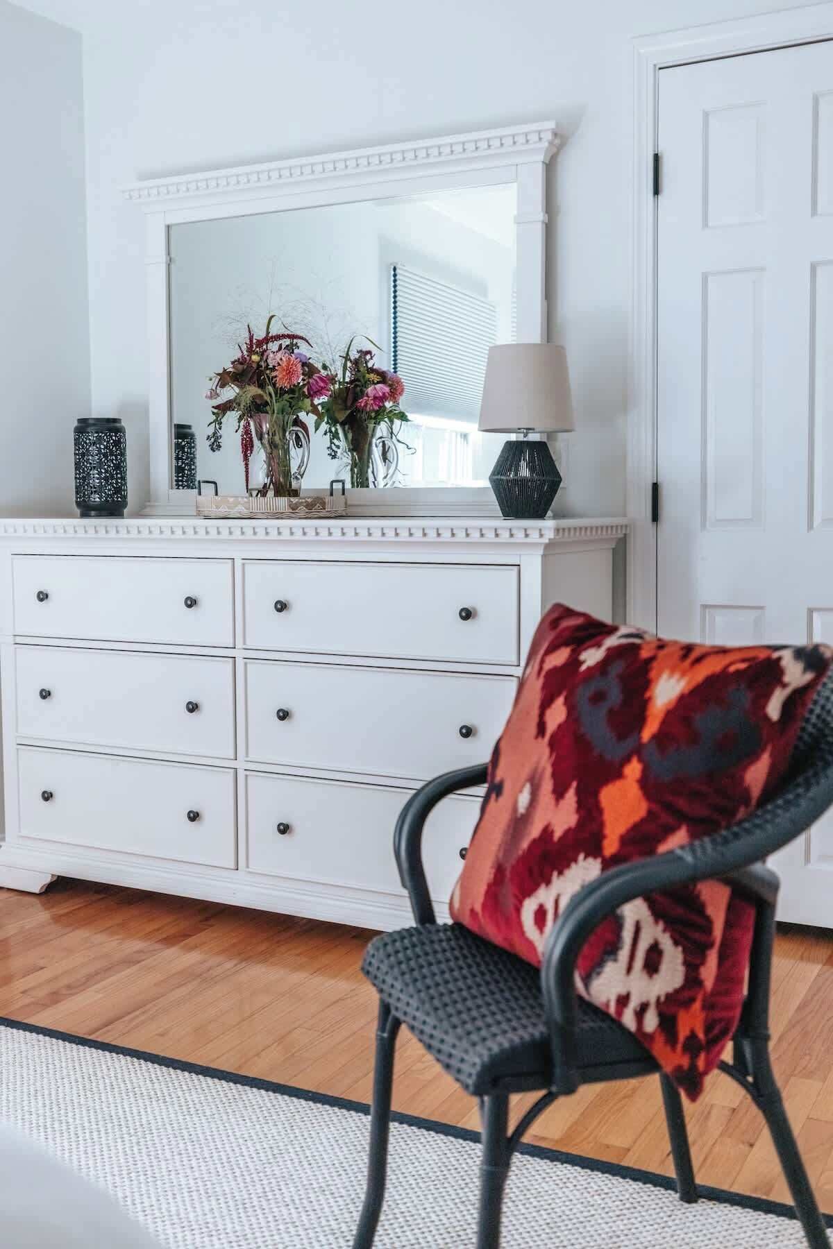 Une commode blanche avec de nombreux tiroirs, un miroir au-dessus, et une chaise avec un coussin à motifs.