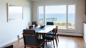 Un comedor con una mesa grande y sillas, vista al mar y un muelle, y un cuadro en la pared.