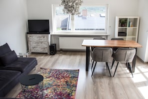Un salon moderne comprenant un canapé, une table basse, une table à manger avec des chaises, un téléviseur posé sur un meuble en bois et une fenêtre donnant sur des panneaux solaires.