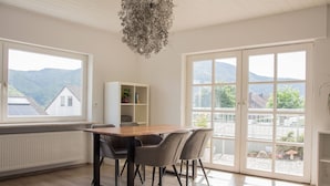 Un coin repas avec une table et des chaises en bois, un meuble blanc orné de plantes et de grandes fenêtres offrant une vue sur les montagnes.