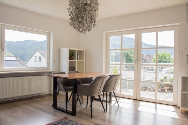 Un coin repas avec une table et des chaises en bois, un meuble blanc orné de plantes et de grandes fenêtres offrant une vue sur les montagnes.