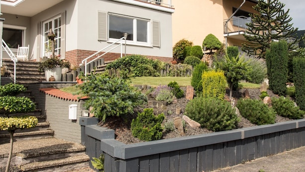 Une maison à deux étages dotée d’un balcon, d’un jardin agrémenté de diverses plantes et arbustes, et d’un chemin de pierre menant à l’entrée.