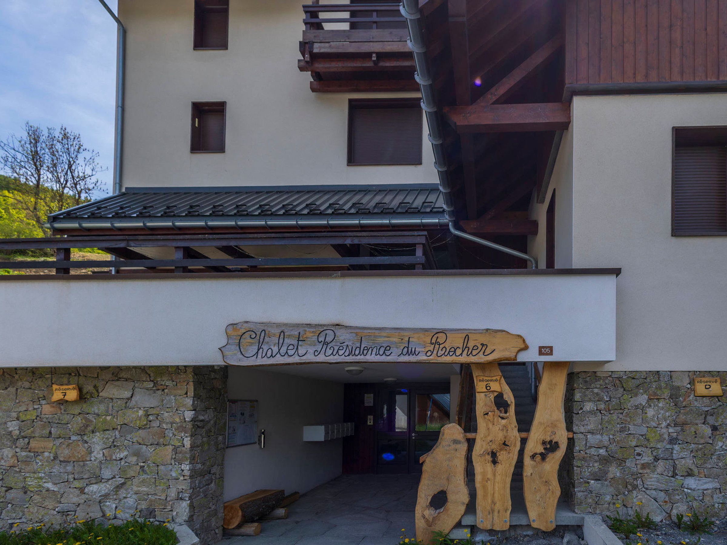 Ein Gebäude mit einem Holunderschild, auf dem 'Chalet Résidence du Raicher' steht, sowie eine Steinmauer mit einer hölzernen Bank.