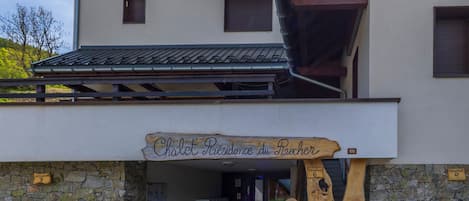 Un bâtiment avec une enseigne en bois indiquant « Chalet Résidence du Raicher » et un mur de pierre sur lequel est adossé un banc en bois.