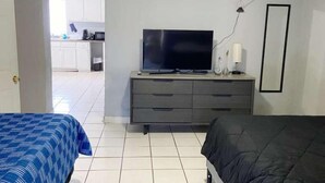 A bedroom with two beds, a TV, a dresser, and a kitchen in the background. - Del Sol Medical Center Gem (El Paso)