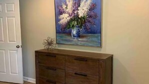 A bedroom with a wooden dresser, a painting of flowers on the wall, and a white door. - Big Blue House Apt 1 (Pikeville)