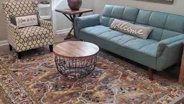 A living room with a patterned armchair, a blue sofa with a 'Welcome' pillow, a round coffee table, and a decorative rug. - Big Blue House Apt 1 (Pikeville)