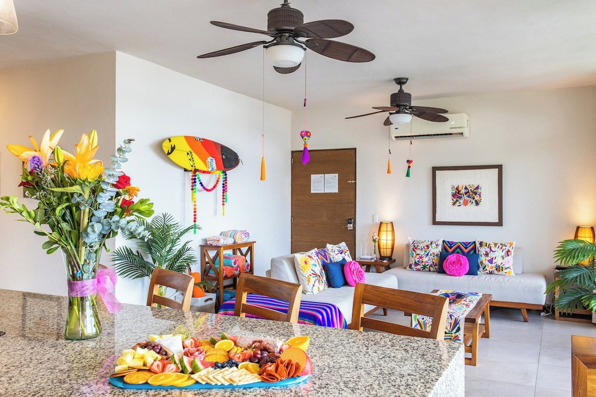 A modern living room with a dining area, featuring a ceiling fan, a sofa with colorful cushions, a coffee table, and a wall-mounted surfboard decoration.