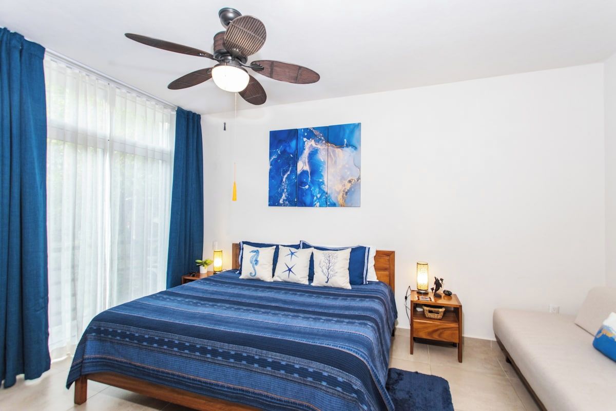 Un dormitorio con cama, mesita de noche, lámpara, ventilador de techo y un cuadro en la pared.