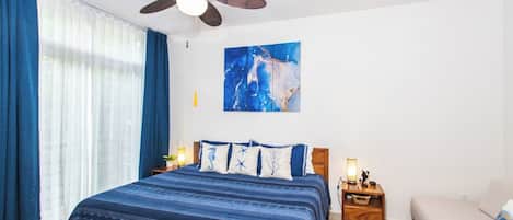 Un dormitorio con cama, mesita de noche, lámpara, ventilador de techo y un cuadro en la pared.