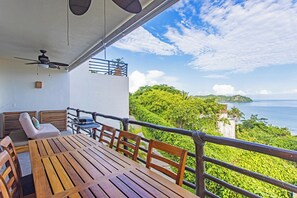 A balcony with a wooden table and chairs, a ceiling fan, and a view of the sea and greenery.