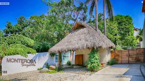 A thatched-roof house with a stone pathway leading to a gated entrance, surrounded by lush greenery and palm trees.
