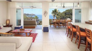 A spacious living area with a sofa, coffee table, and dining area with chairs and a table. There is a large window offering a view of the sea and greenery.