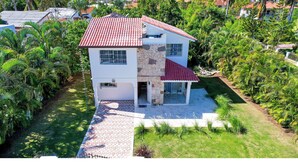 Une maison à deux étages, avec un toit recouvert de tuiles rouges, entourée d’une végétation luxuriante et de palmiers.