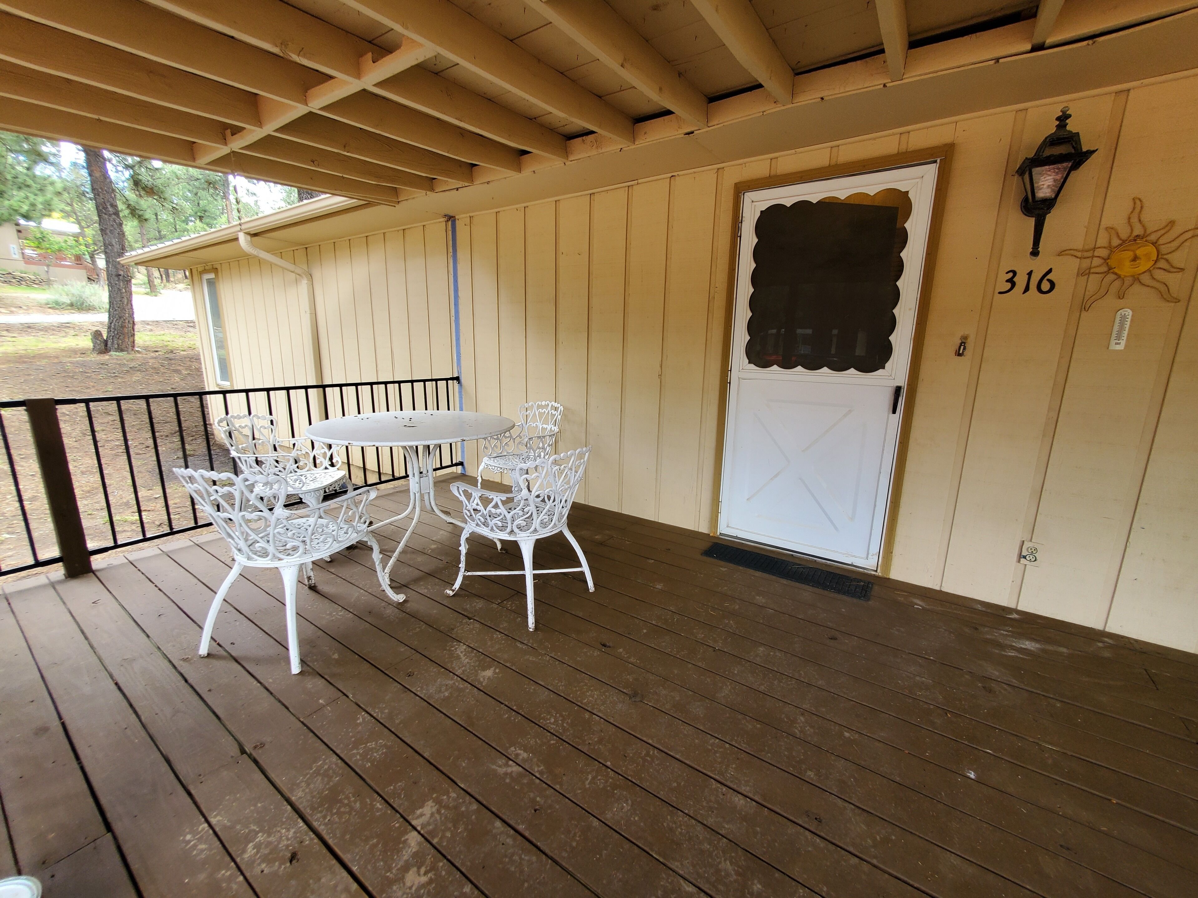 Un porche abrité avec une terrasse en bois, une porte blanche portant un numéro et une petite table et des chaises d’extérieur.