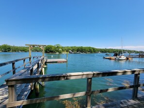 A wooden pier extending into a calm body of water with boats docked nearby and a forested shoreline in the background.