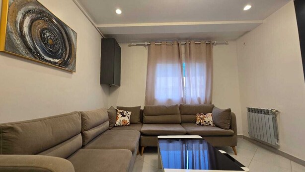 Un salon comprenant un canapé d’angle, une table basse, un tableau accroché au mur et une fenêtre avec des rideaux.