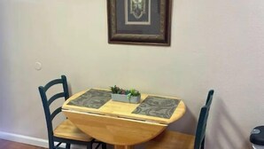 A small dining table with two chairs, a framed picture on the wall, and a trash can to the side. - The   Nook (Pikeville)