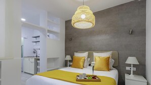 A modern hotel room with a bed, bedside tables, a hanging light fixture, and a view into the kitchen area.
