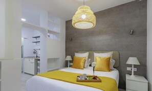 A modern hotel room with a bed, bedside tables, a hanging light fixture, and a view into the kitchen area.