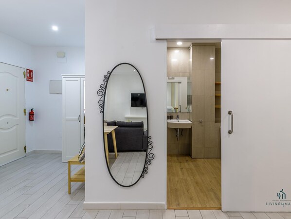 A hotel room with a large mirror, a sofa, a small table, and a bathroom with a sink and mirror.