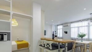 A modern hotel room with a bed, a dining table, chairs, a microwave, and an air conditioning unit.