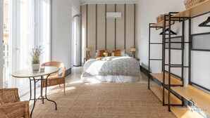 Un dormitorio con una cama, una mesita redonda pequeña, sillas de mimbre, un banco de madera y un estante en la pared.