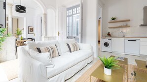 Una sala de estar moderna con un sofá blanco, una mesa de centro y una zona de cocina con armarios blancos y una lavadora.