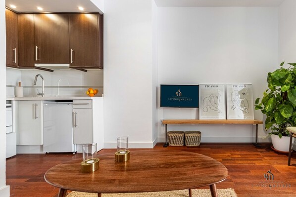 Una cocina moderna con armarios de madera, encimera blanca y comedor con una mesa redonda. Se observan obras de arte enmarcadas en la pared y una planta en maceta en la esquina.