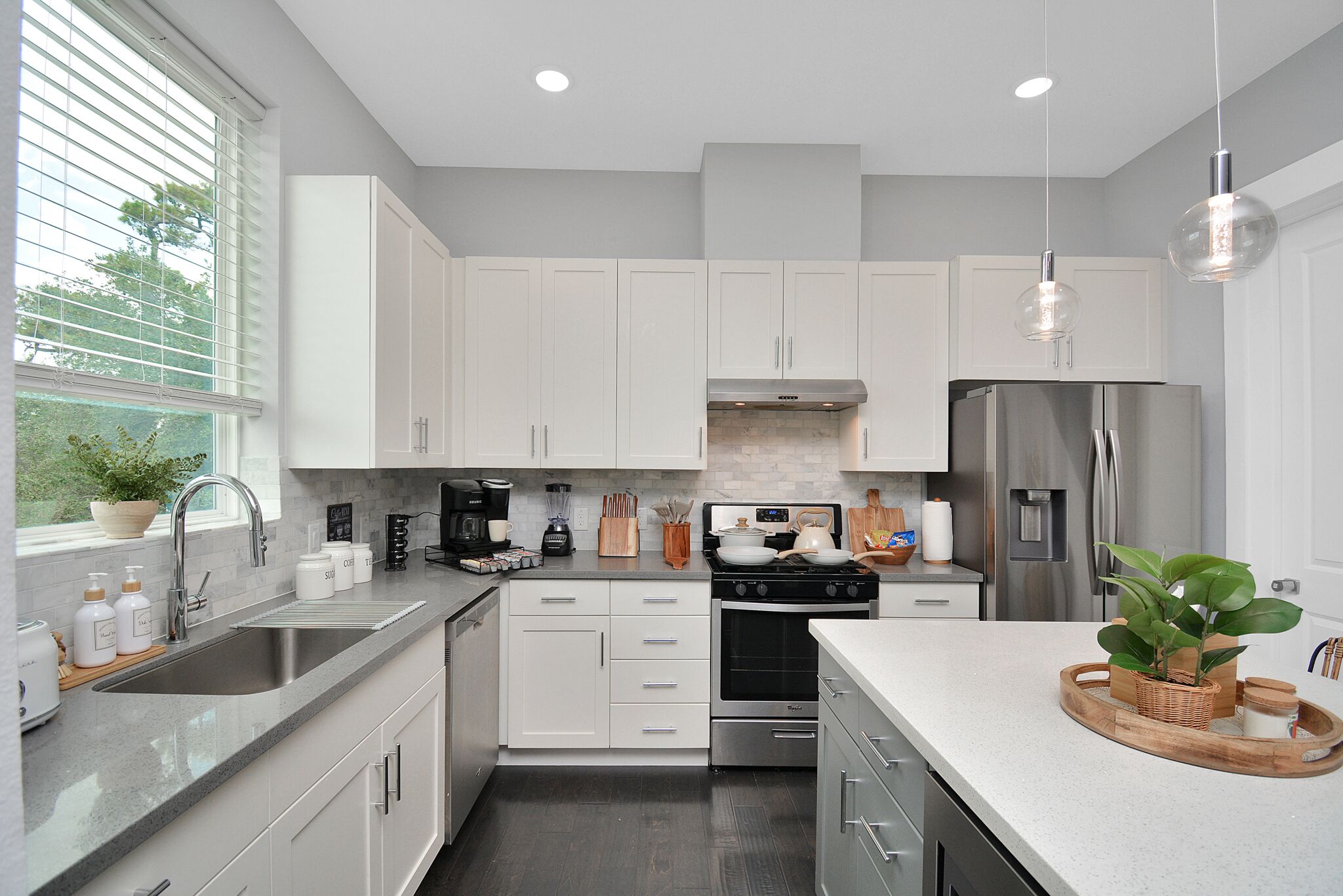 A modern kitchen with white cabinets, stainless steel appliances, and a large window with blinds.