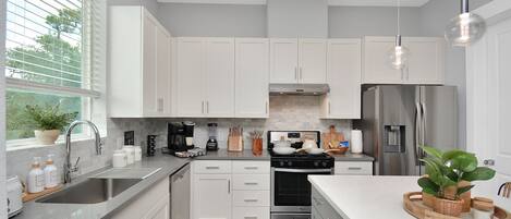 A modern kitchen with white cabinets, stainless steel appliances, and a large window with blinds.