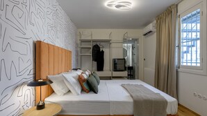 A modern bedroom with a wooden headboard, a bedside table with a lamp, a bed with multiple pillows, a wardrobe, and a window with curtains.