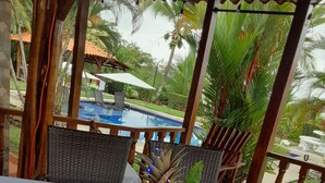 A table set with a tropical-themed tablecloth, a pineapple, coconuts, and decorative elements in a covered outdoor area with a pool in the background. - Cozy Iguana Apartmentos 3 (Esterillos Oeste)