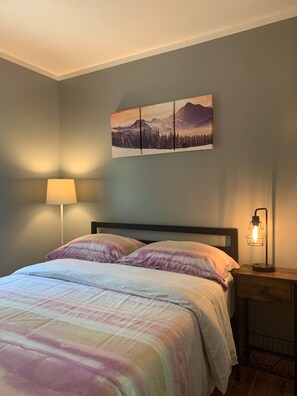 Un dormitorio con cama, mesita de noche, lámpara y un cuadro colgado en la pared.