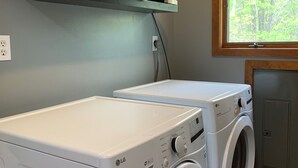Een wasserette met twee LG DirectWave wasmachines, een plank met opgevouwen handdoeken, een raam met uitzicht op bomen en een grijs kastje.