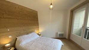 A bedroom with a wooden headboard, a bed with white bedding, bedside lamps, a small trash bin, and a window with blinds. - Veronica n°53: T2 with mountain corner (Châtel)
