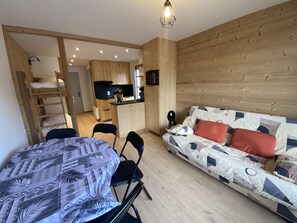A compact living space with a bunk bed, a kitchenette, a dining area with a table and chairs, and a sofa with patterned cushions.