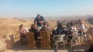 Eine Gruppe von Personen sitzt auf Plastikstühlen an einer erhöhten Aussichtslage und blickt auf eine weite Landschaft.